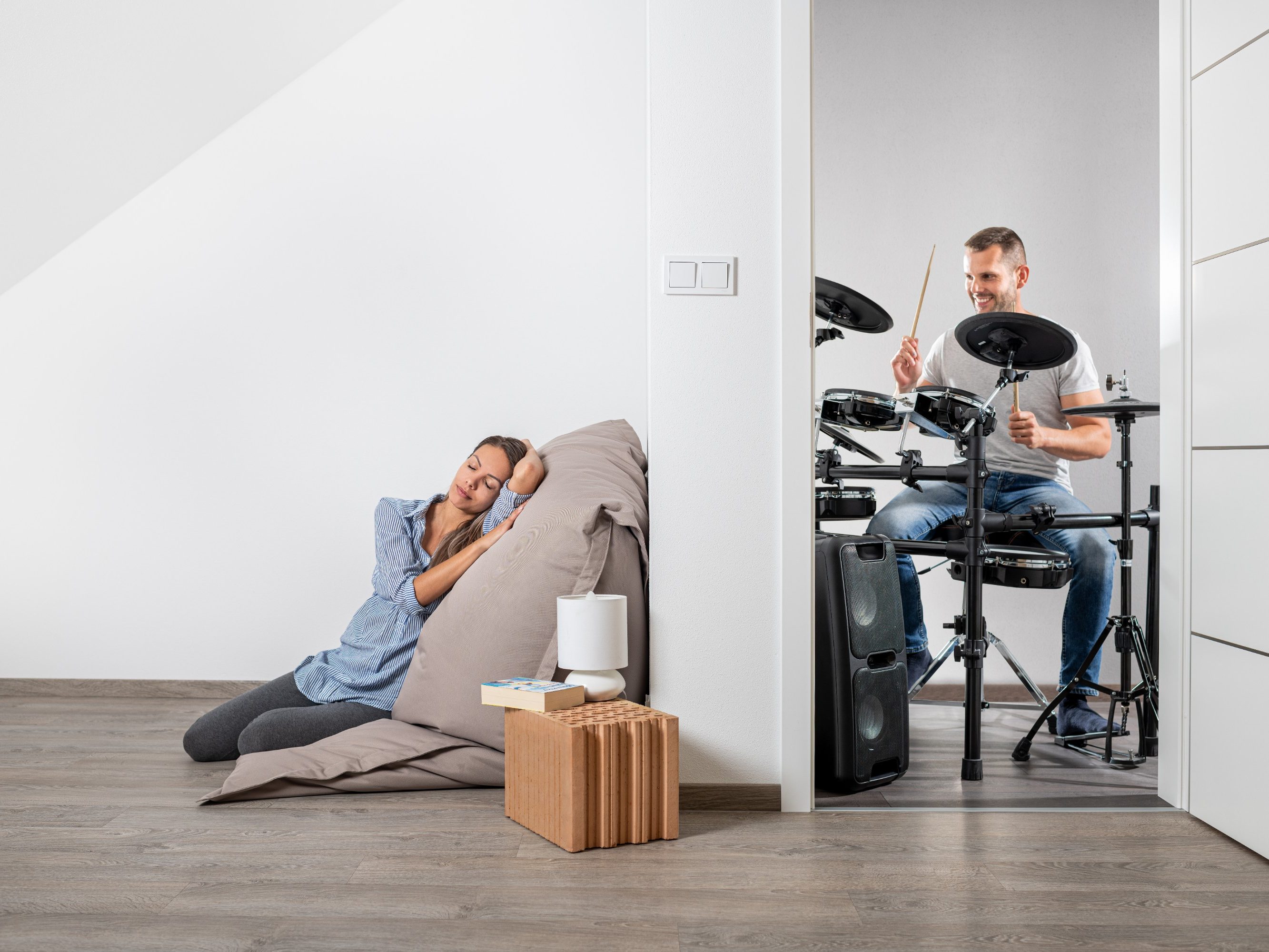 Žena v klidu ležící na polštáři v jedné místnosti přičemž muž v druhé místnosti hraje na bicí | Zdravý domov