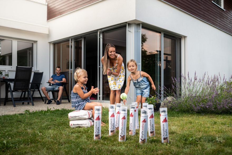 Děti si hrají před domem na zahradě s maminkou na kuželky se zdicí pěnou Porotherm Dryfix a lepidlem Porotherm Dryfix.extra